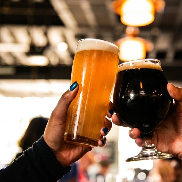 Two hands clinking glasses of light and dark beer in a lively restaurant setting.