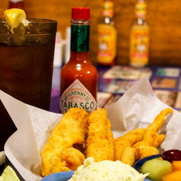 Fried fish basket with coleslaw, fruit, iced tea, and Tabasco sauce on a colorful table.