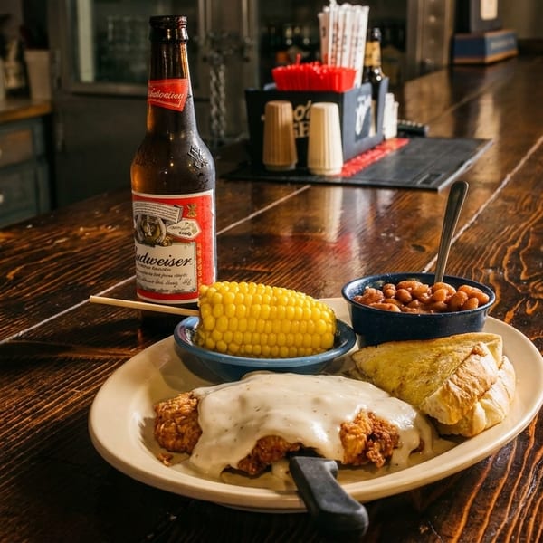 Chicken fried steak with gravy, corn, baked beans, toast, and a beer on a rustic wooden bar.